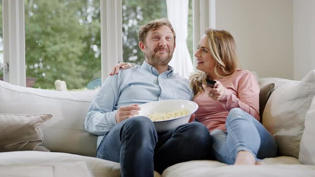 Couple On Date Night Sitting On Sofa At Home Laughing And Watching Movie On TV With Popcorn Together - Shot In Slow Motion