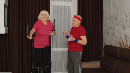 Old senior couple grandfather and grandmother exercising on orbitrek, doing weight lifting sport dumbbells exercises in room at home. Retro style Caucasian mature elderly man and woman warming up