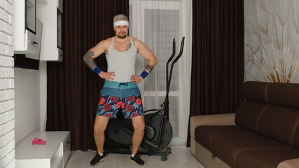 Fototapeta na wymiar Funny man athlete sportsman guy from the 80's with beard makes workout stretching at home. Portrait of young stylish hipster man practicing sport, having fun, fooling around. Fitness parody concept