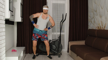 Fototapeta na wymiar Funny man athlete sportsman guy from the 80's with beard makes workout stretching at home. Portrait of young stylish hipster man practicing sport, having fun, fooling around. Fitness parody concept