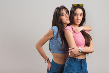 Young twin sisters hugging. Pretty teen twins wearing striped t-shirts and jean shorts on light grey background