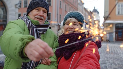 Senior old couple with burning sparklers celebrating. Elderly family holding bengal lights enjoying Christmas eve on city street during coronavirus lockdown. Active pensioners celebrating birthday