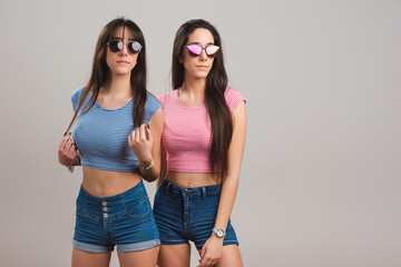 Young twins. Pretty teen twin sisters wearing sunglasses, striped t-shirts and jean shorts on light grey background