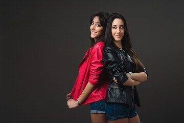 Young twins. Twin sisters standing back to back wearing leather jackets on dark grey background