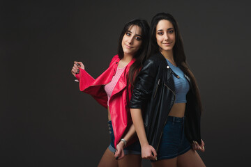 Young twins. Twin sisters standing back to back wearing leather jackets on dark grey background