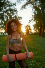 Naklejka na ściany i meble Vertical shot of a young happy mixed race woman wearing sport clothes holding yoga mat and smiling at camera while standing in park in the early morning, exercising outdoors
