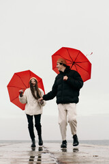 Young hipster couple walking with two red umbrellas in cold weather, love story outdoors