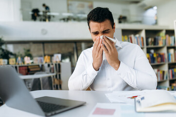 Unhealthy male employee sit at the desk, using a paper tissue, sneezing. Business man feeling unwell, struggling with running nose. Hispanic guy remotely working or studying, while sickness