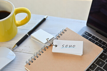 There's a word book on a desk with a cup of coffee and laptop pc. The word SOHO is written in it. It's an abbreviation for Small Office Home Office.