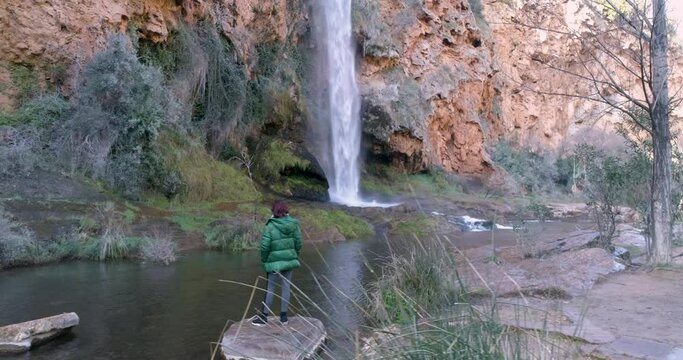 Mujer de espalda con abrigo verde contemplando ca&iacute;da de agua vista a&eacute;rea de dron