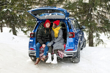 Romantic young hipster couple hugging while sitting in car trunk under falling snow. Love, valentines and holiday concept. Kiss and hug. Valentines day celebration and happiness concept