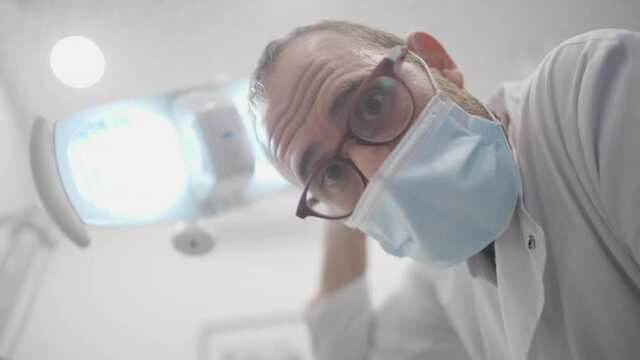 Male dental doctor looking down at patient while moving the led dental lights around trying to focus the light. White male orthodontist before starting procedure. POV