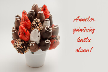 Happy mother's day greeting card that reads Anneler gununuz kutlu olsun with red lettering; A bundle of edible flowers, arrangement of strawberries covered with chocolate isolated on white background