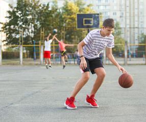 Young cute boy plays basketball at the outdoor streetball court on a sunny summer day. Teenager player in action dribbling the ball. Hobby, active lifestyle, sports for kids.