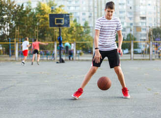Fototapeta premium Young cute boy plays basketball at the outdoor streetball court on a sunny summer day. Teenager player in action dribbling the ball. Hobby, active lifestyle, sports for kids.