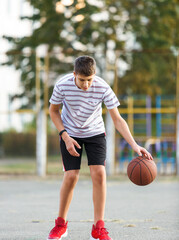 Naklejka premium Young cute boy plays basketball at the outdoor streetball court on a sunny summer day. Teenager player in action dribbling the ball. Hobby, active lifestyle, sports for kids.
