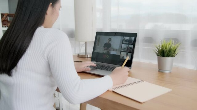 Online classroom video call with classmates and professor teaching educational learning, social distancing working from home workspace, happy smiling Asian girl writing on note book revising studying