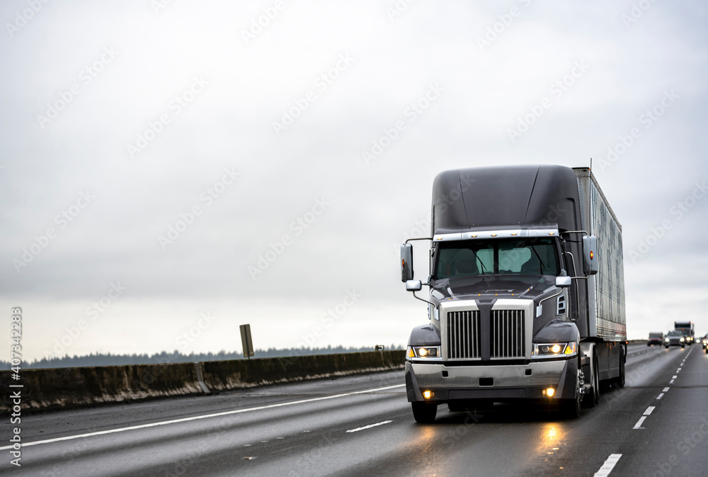 Wall mural stylish dark gray big rig semi truck with turned on headlights running with semi trailer on the stra