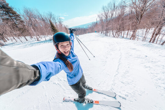 Ski selfie happy skier woman screaming of joy skiing on ski resort slopes with arms up in fun. Asian girl wearing winter jacket, helmet, gloves, boots and skis.