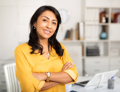Happy smiling Latin American businesswoman standing in office with arms crossed looking at camera