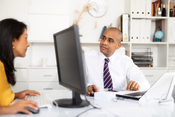 Fototapeta premium Portrait of positive hispanic office employee during daily work with computer, sitting at desk with woman colleague