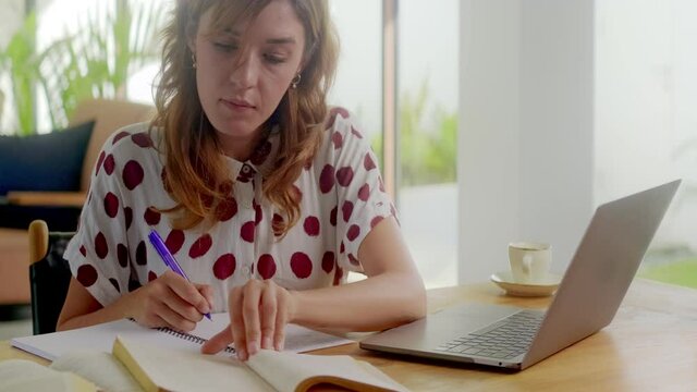 intelligent millennial female student studying on laptop writing notes, serious young woman prepare for test exam doing research project coursework homework learning at home using computer at desk
