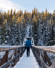 Man in a blue jacket, black pants walking across a snowy, snow covered bridge in winter time with boreal forest and winter skies, blue sky in background in Miles Canyon, Yukon. 