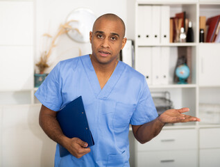Latin american doctor wear blue medical uniform, stethoscope and clipboard look at camera posing in private clinic