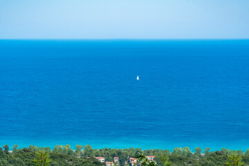 In far distance a single floating yacht with open white sails traveling in tranquil waters. Turquoise blue sea and sky background with copy space. Green trees and houses on bottom frame