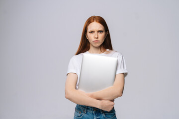 Distressed upset young business woman or student holding laptop computer and looking at camera on isolated gray background. Pretty lady model with red hair emotionally showing facial expressions.
