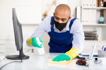 Man in protective mask and in an overalls cleans the table at office