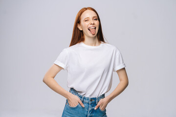 Joyful young woman wearing T-shirt and denim pants showing tongue on isolated white background. Pretty lady model with red hair emotionally showing facial expressions in studio, copyspace.