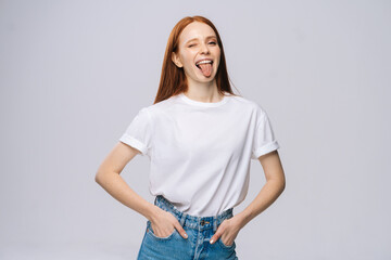 Obraz premium Joyful young woman wearing T-shirt and denim pants showing tongue on isolated white background. Pretty lady model with red hair emotionally showing facial expressions in studio, copyspace.