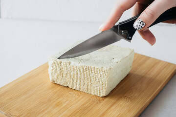metal knife in hand cuts large piece of tofu cheese on wooden desk on white background. Healthy diet vegetarian food,selective focus