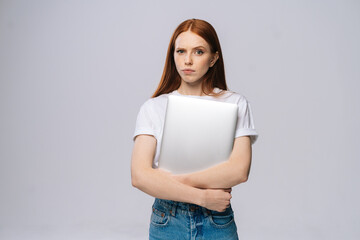 Frustrated upset young business woman or student holding laptop computer and looking at camera on isolated gray background. Pretty lady model with red hair emotionally showing facial expressions.