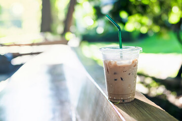 Iced coffee in a plastic cup with a straw placed on a wooden table surface blurred green...