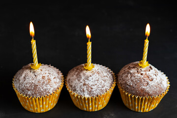 Three cupcakes in paper cups with yellow candles with flame surprise birthday on dark background