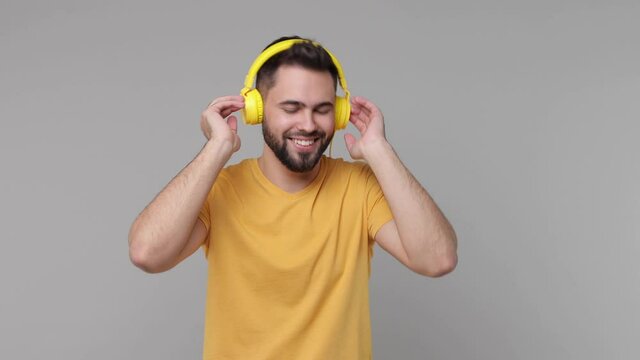 Cheerful smiling bearded young man 20s years old in yellow casual t-shirt isolated on grey color background studio. People lifestyle concept. Listening music with headphones dancing clenching fists