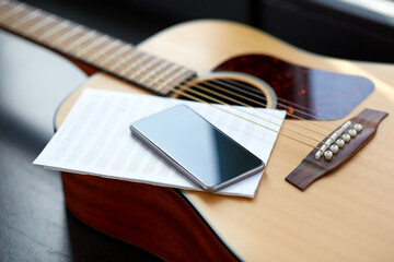 technology and music writing concept - close up of acoustic guitar, music book and smartphone