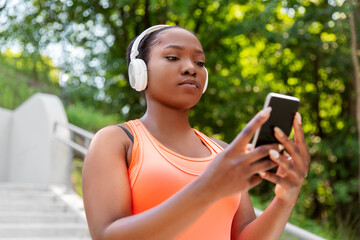 fitness, sport and technology concept - young african american woman with headphones listening to music on smartphone outdoors