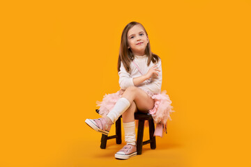 Young girl in tulle skirt and white pullover sitting on small wooden stool on bright yellow background - Powered by Adobe