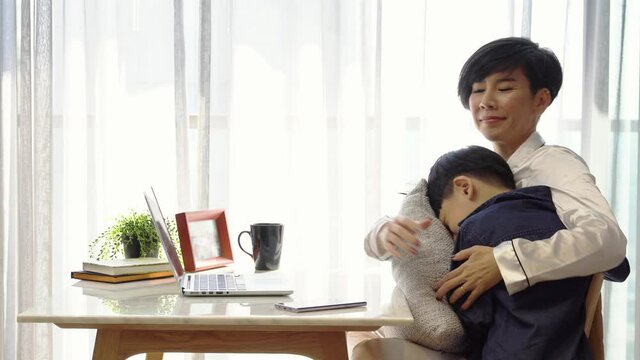 Beautiful Asian mother work at home, typing on computer laptop. Boy child walk to her to hug and kiss. Technology, new normal, social distancing, work life balance during Covid 19 pandemic concept.
