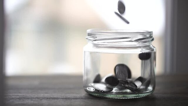 Ukrainian coins falling into the glass jar on wooden table near window, close up. Slow motion, a pile of silver coins falls into an empty glass jar on a window background. Ukraine
