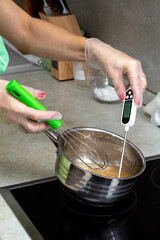 Gloved hands hold a pot of syrup, a whisk, and a thermometer. The cooking process. Selective focus.