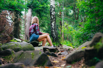 Hipster girl in the mountains. Stylish woman in checkered shirt sitting on stones in forest. Wanderlust concept. Hiking and travelling in summer. Mysterious wood. Beautiful nature.
