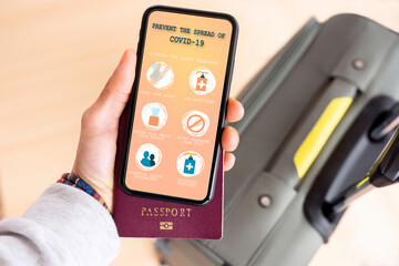 (Selective focus) Overhead view of an human hand holding a passport and a smart phone with a digital illustration of some guidelines to prevent the spread of Covid-19 disease.