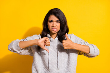 Photo of charming unsure dark skin girl dressed grey sweatshirt cannot choose thumb up down...