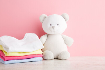 Stack of colorful baby clothes and white teddy bear on wooden table at light pink wall background. Pastel color. Closeup. Front view.