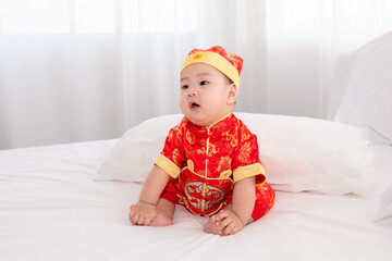 Adorable Asian boy in red Chinese Costume clothes sitting on bed at home looking for somethin with curiosity, innocent cute baby feel sleepy yawning boring waiting for mother feeding