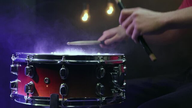 A man plays with sticks on a snare drum with dust in a dark room with a beautiful light.